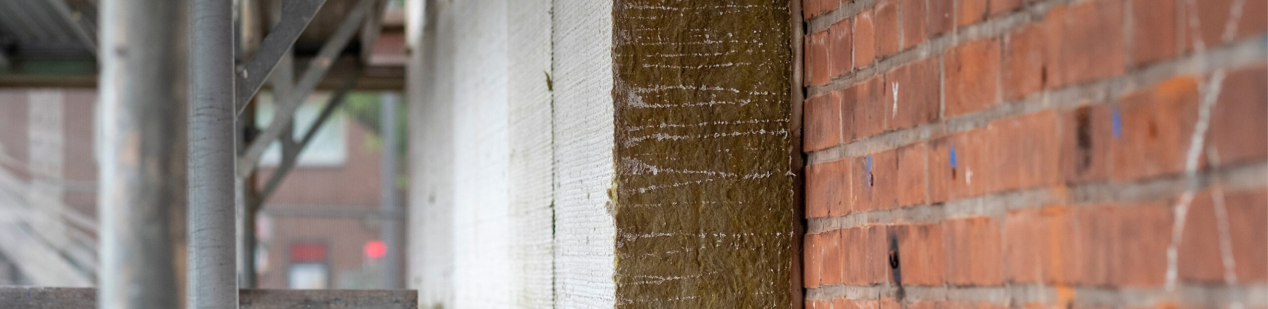 Eine Nahaufnahme eines Bauprojekts, die den Schichtaufbau einer Außenwand zeigt, von links nach rechts bestehend aus metallischem Baugerüst, einer hellen Dämmplatte und einer freiliegenden, rauen Dämmschicht vor der originalen roten Backsteinwand.