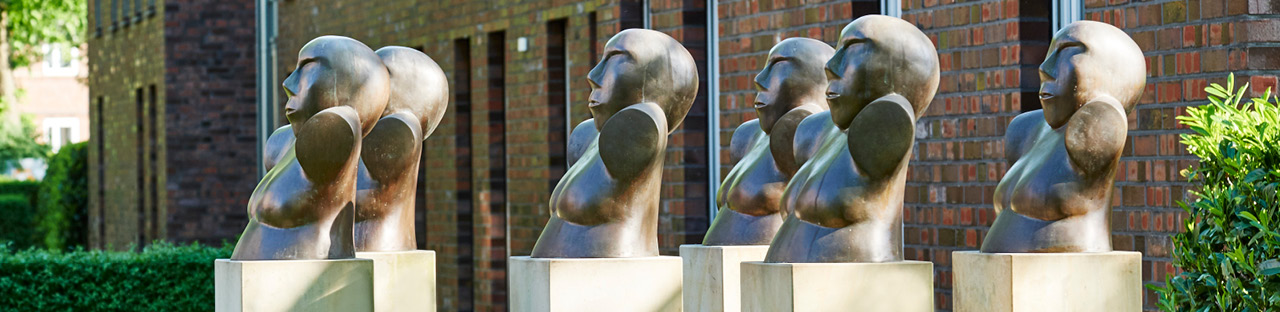 Eine Reihe von sechs bronzefarbenen, abstrakten Büsten in Seitenansicht, die auf hellen Podesten vor einer Fassade aus rotem Backstein und großen Fenstern stehen.