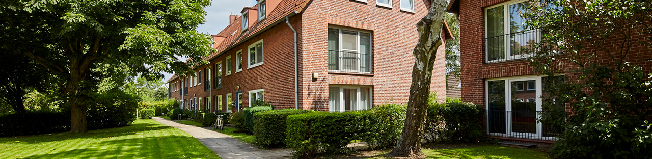 Eine Reihe von traditionellen Backsteinhäusern in einer grünen Wohngegend. Die Häuser haben weiße Fensterrahmen und einige kleine Balkone. Im Vordergrund sind gepflegte Rasenflächen, Büsche und Bäume zu sehen, mit einem schmalen Fußweg, der sich durch die Szene schlängelt. Das Sonnenlicht fällt auf die Gebäude und das Grün.